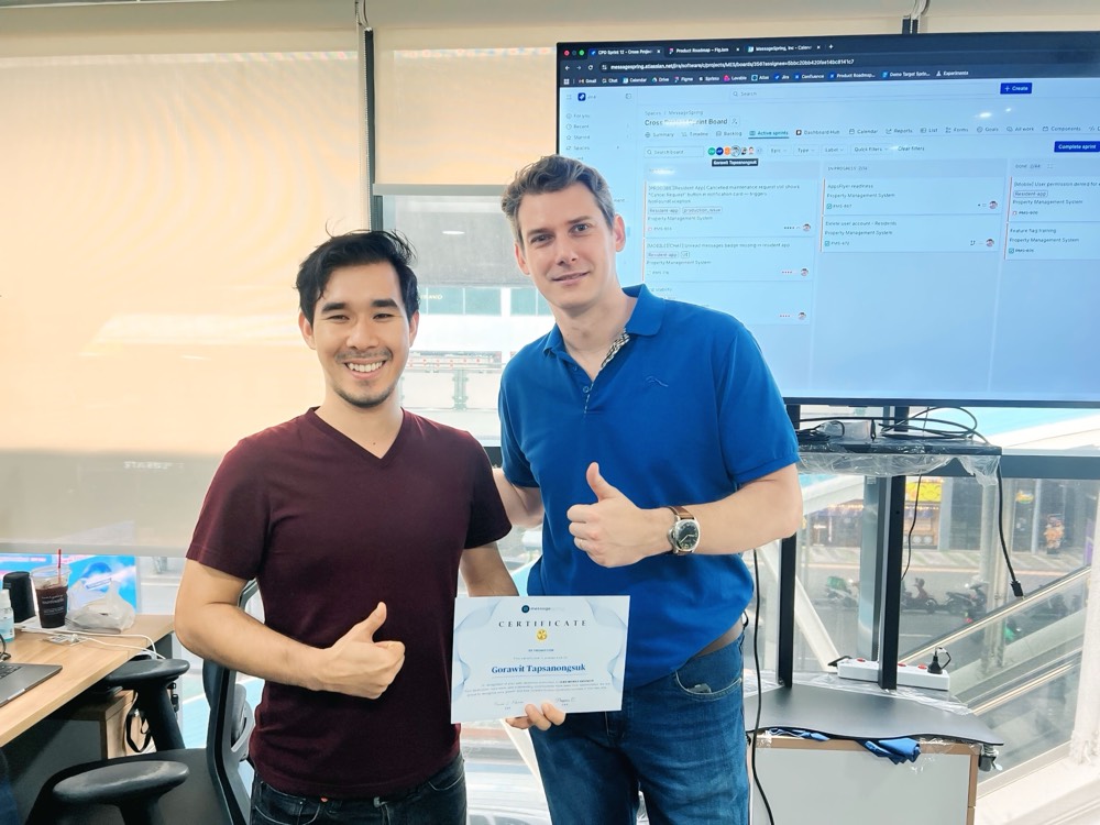 A teammate holding his promotion certificate beside a colleague giving a thumbs-up, the sprint board on screen behind them